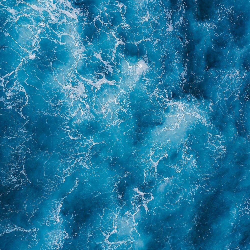 Overhead view of bright turquoise ocean water with white wave patterns and foam textures creating an abstract, dynamic sur...