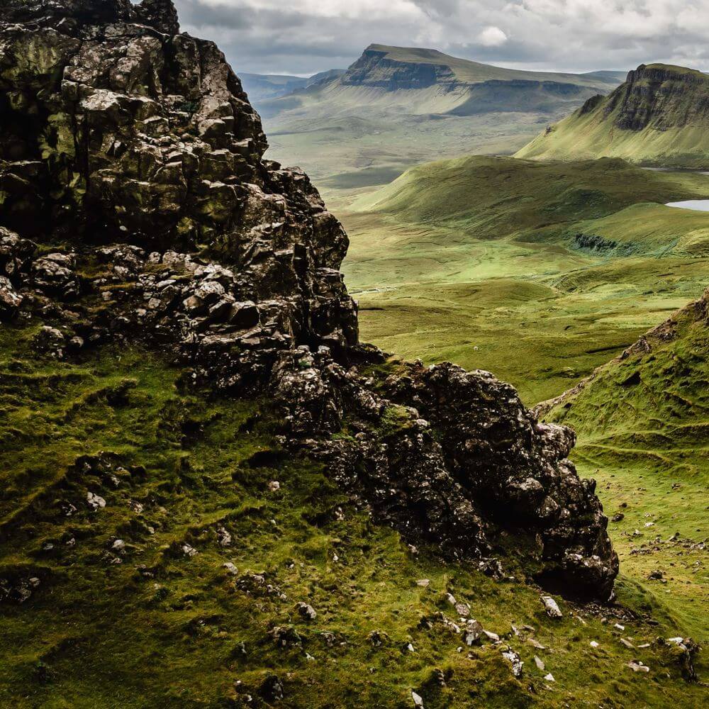 Scottish Highland landscape with moss-covered rocky outcrops, rolling green valleys, distant mountains, and small lochs un...