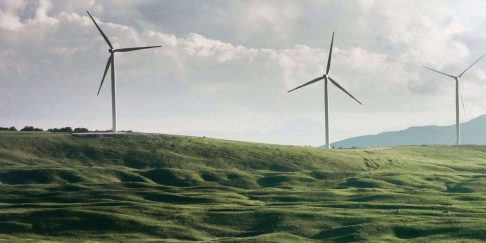 Wind turbines in rolling green hills with mountains in the distance under cloudy sky.