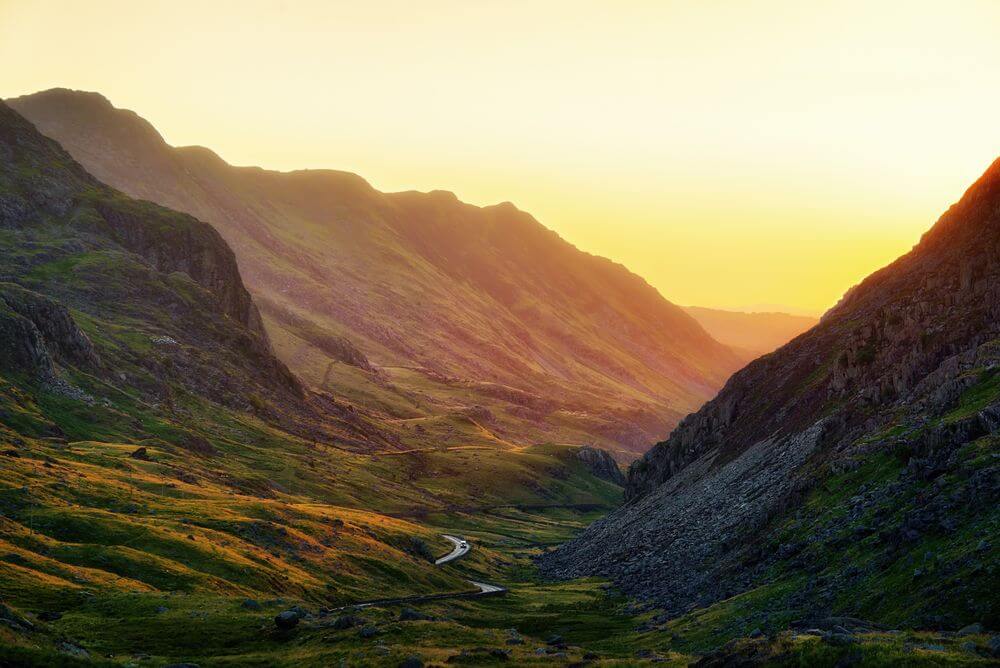Scenic mountain valley with winding river at golden hour sunset, green slopes and dramatic peaks under warm light.