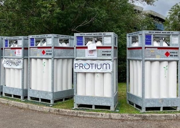 Industrial storage containers labeled "PROTIUM" on production set, surrounded by green trees and equipment.