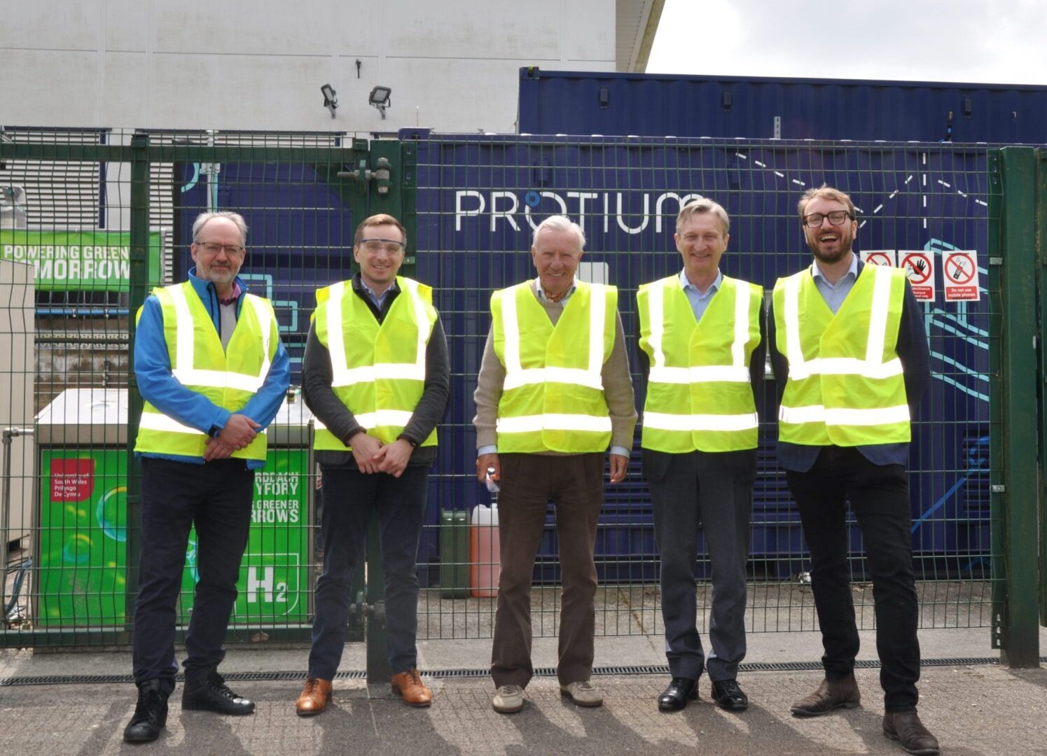 Five people wearing safety vests stand in front of a blue Protium hydrogen facility container at a Welsh industrial site.