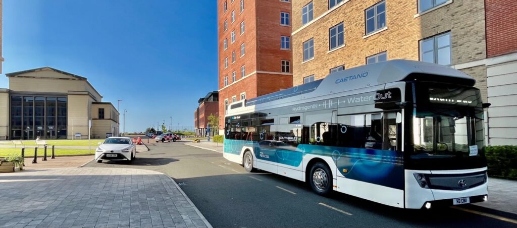 Hydrogen fuel cell bus with blue and white livery parked in urban area near brick buildings during trial operations.