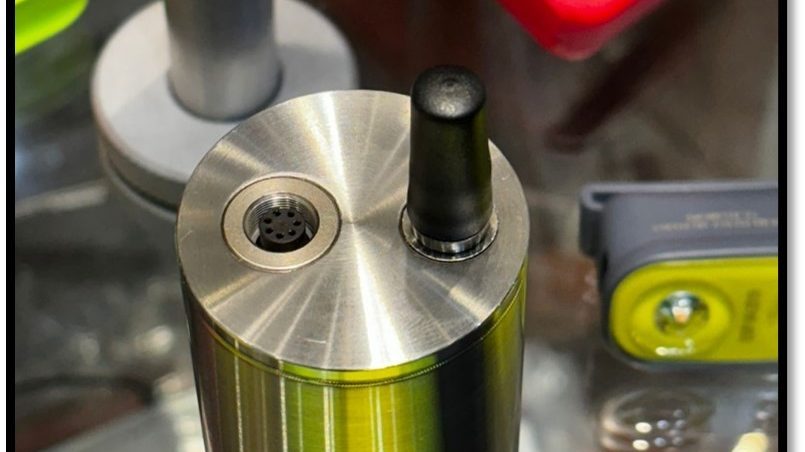 Close-up of a vaping device with green and black coloring, showing the metal top cap with airflow holes and mouthpiece, po...