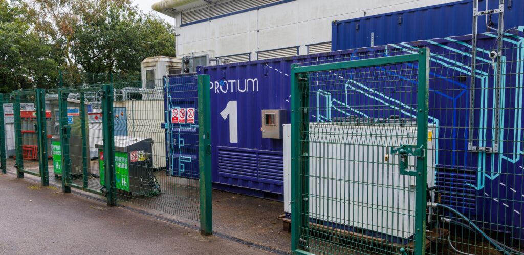 Protium Pioneer-1 facility exterior with blue container marked "PROTIUM 1" behind green security fencing.