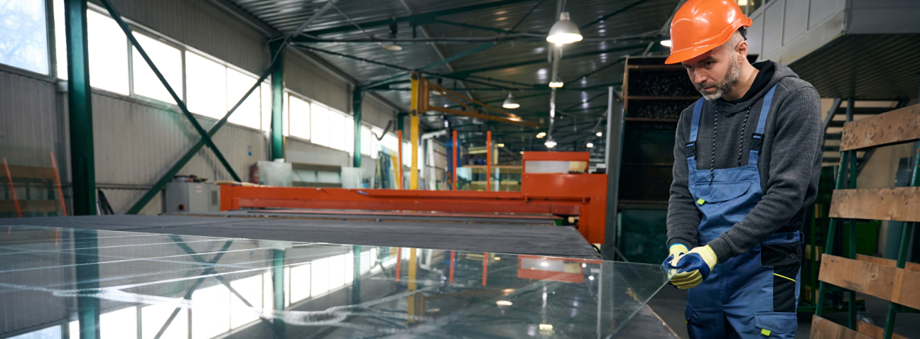 Worker in orange hard hat and blue overalls inspecting steel sheet in industrial factory warehouse with machinery and meta...