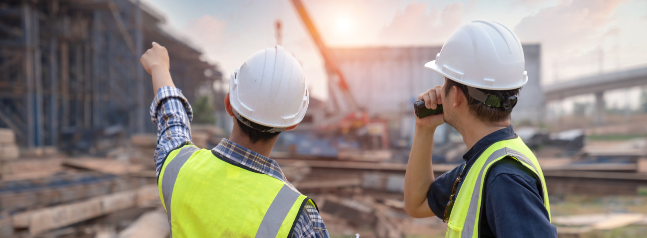 Two construction workers in hard hats and safety vests observe a busy industrial site with machinery and buildings at sunset.