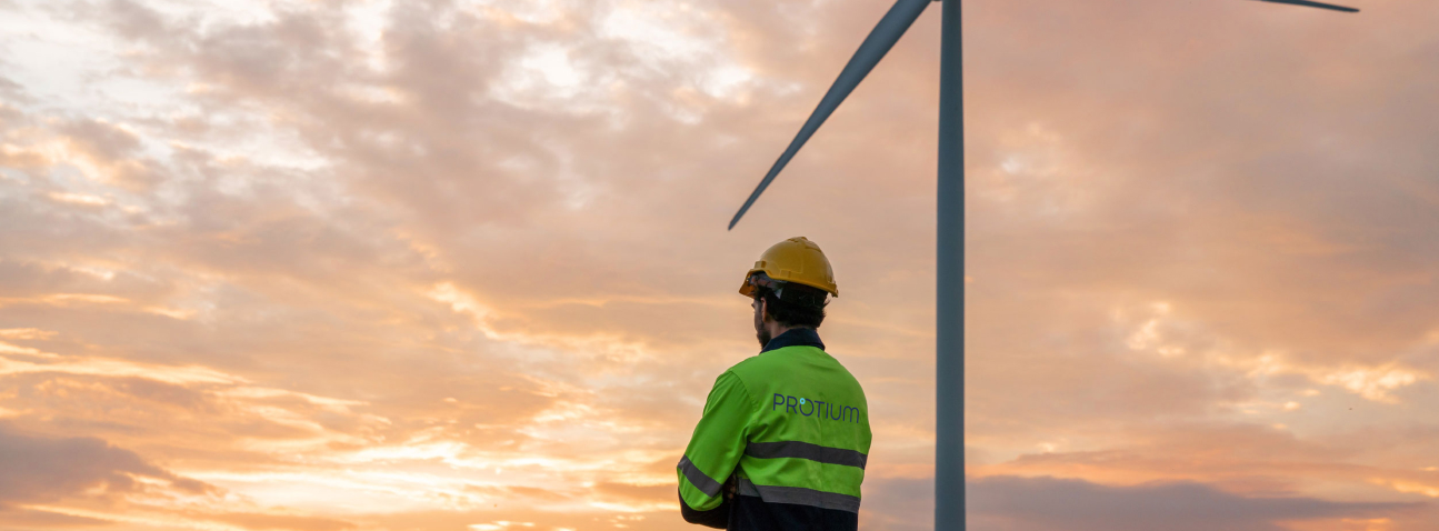 Construction worker in safety gear observing wind turbine against golden sunset sky during commercial development project.