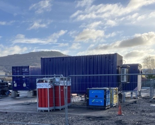 Protium hydrogen generator unit with red and blue containers at construction site, mountains visible in background.